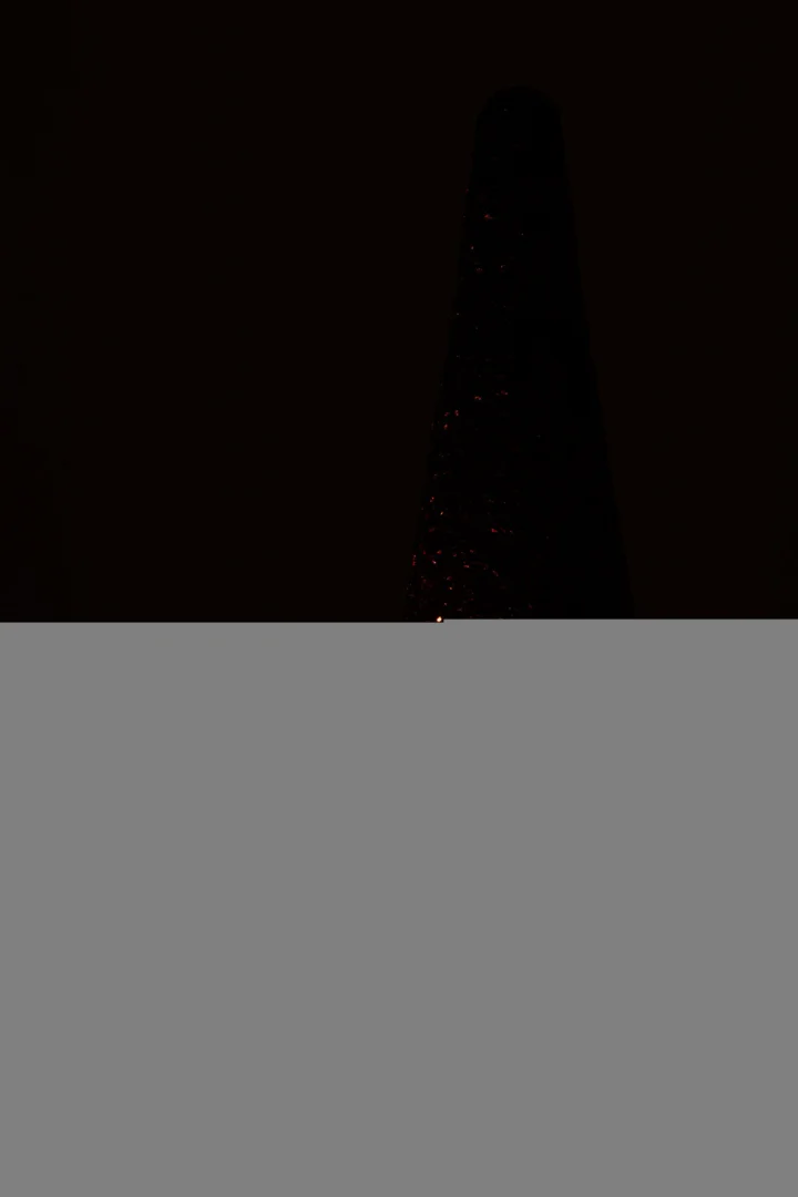 De Theelichthouder Glitter Glas Rood heeft een hoog, donker ontwerp met verspreide glinsterende lichtjes, dat afsteekt tegen een zwarte achtergrond en 's nachts op een gloeiende toren of kegel lijkt.