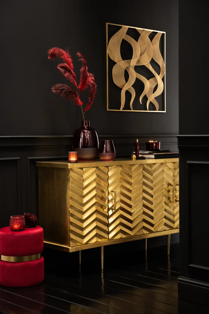 Een gouden kast met chevronpatroon staat tegen een donkere muur met daarop Theelichthouder Glitter Glas Rood vazen, bordeauxrode kaarsen en rood pampagras. Erboven hangt gouden abstracte kunst. Vlakbij staat een rode poef op een zwarte vloer.