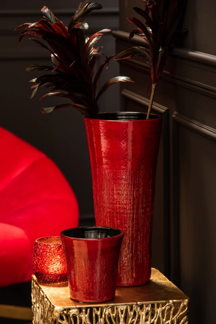 Twee glanzend rode vazen - een hoge met donker gebladerte en een kleine - staan op een goudkleurige tafel met textuur. Daarnaast staat de Theelichthouder Glitter Glas Rood kandelaar. Op de donkergekleurde achtergrond staat een rode stoel.