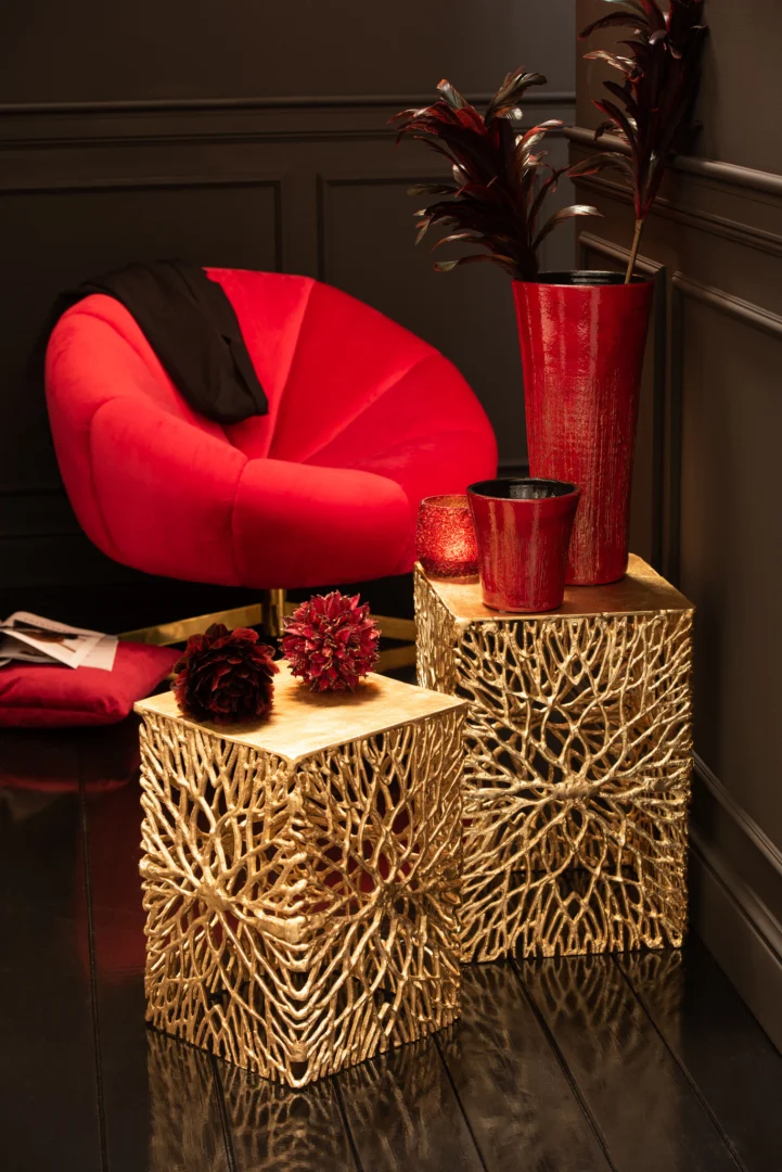 Een stijlvolle kamer met Theelichthouder Glitter Glas Rood, gecombineerd met een rood fluwelen stoel, gouden kubustafels en donkerrode bladvazen tegen donkere wanden met panelen en glanzend zwarte vloeren.