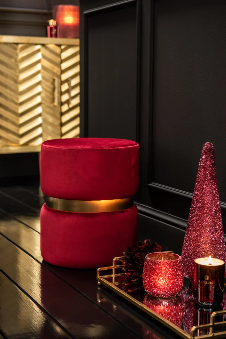 Een rood fluwelen poef met een gouden band ligt op een donkere houten vloer naast een gouden dienblad met Theelichthouder Glitter Glas Rood, een dennenappel en een glitterdecoratie van rode kegels, met een gouden kast op de achtergrond.