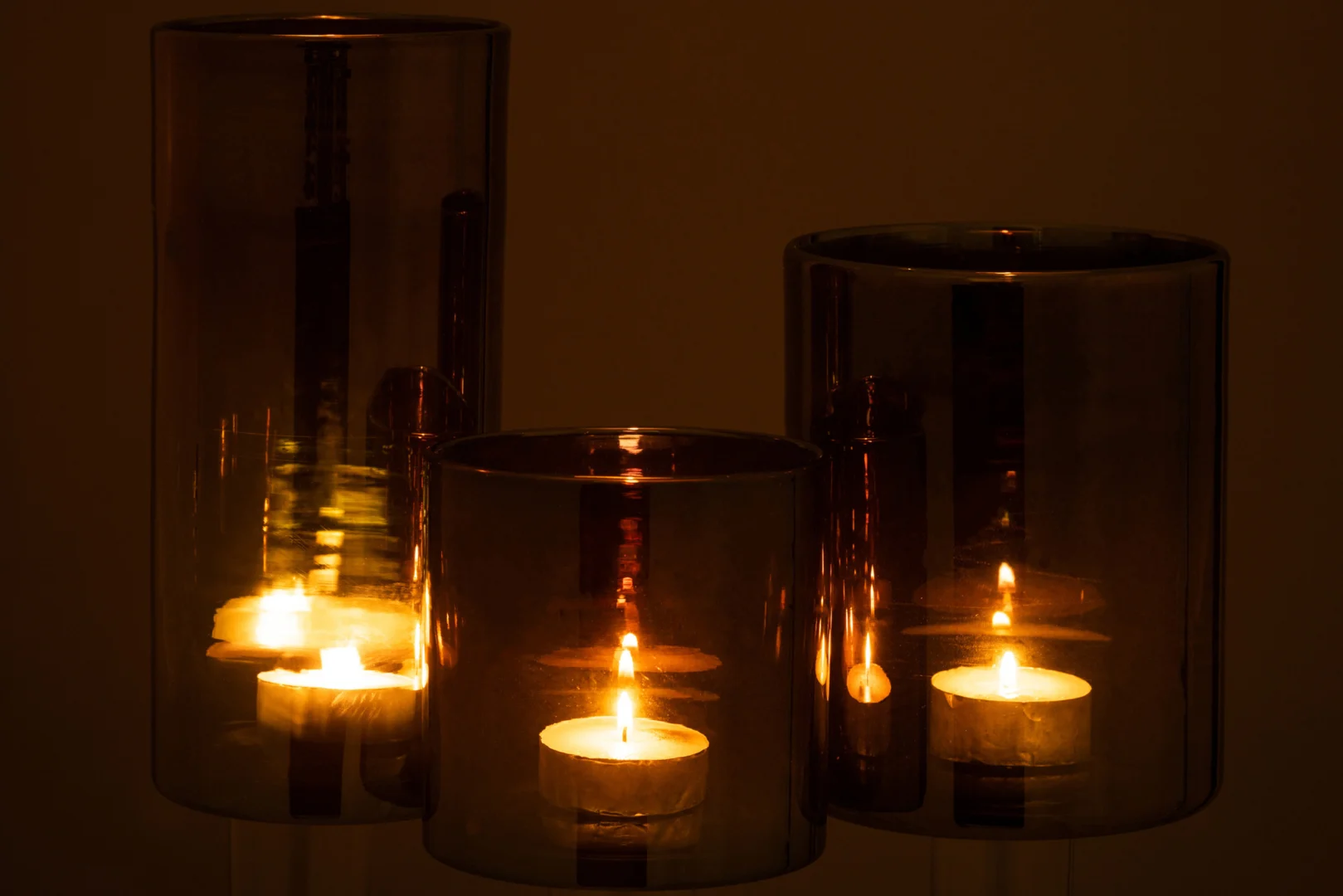 Drie waxinelichtjes in de Kaarsenhouder Hurricane Glas Donkergrijs Groot creëren een warme, zachte gloed door het donkergrijze cilindervormige glas en versterken de sfeer van een schemerige ruimte.