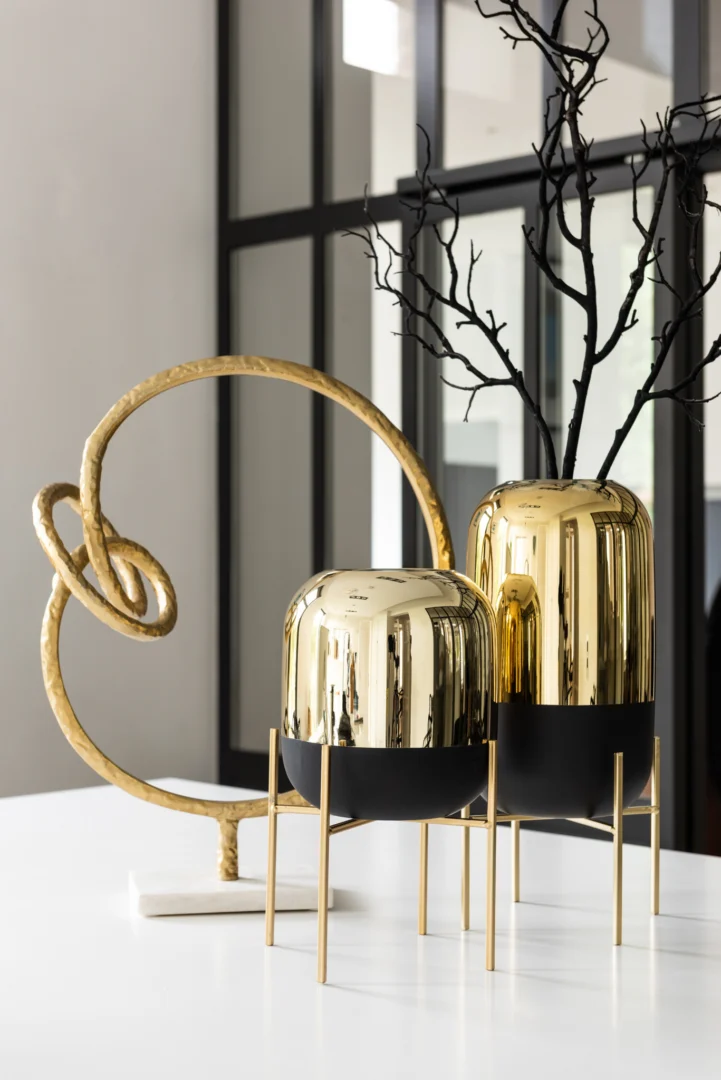 De Kaarsenhouder Orkaan Te Voet Glas Zwart Goud Klein staat op dunne poten met kale takken naast een sculpturaal ornament met gouden lussen op een witte tafel, tegen grote ramen met zwarte omlijsting.