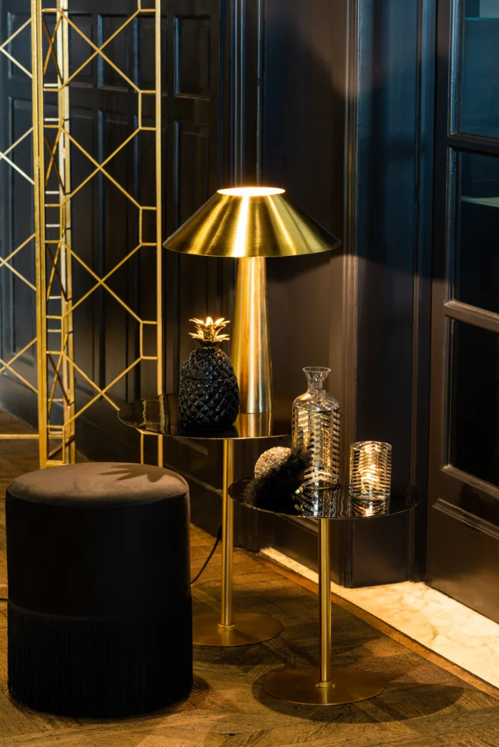 Een luxueus interieur met tafels met goudaccenten en de Kaarsenhouder Orkaan Streep Glas Goud Transparant Klein, glasdecor, een zwart ananasornament en een zwarte voetenbank tegen een wand met donkere panelen en een vloer van visgraathout.
