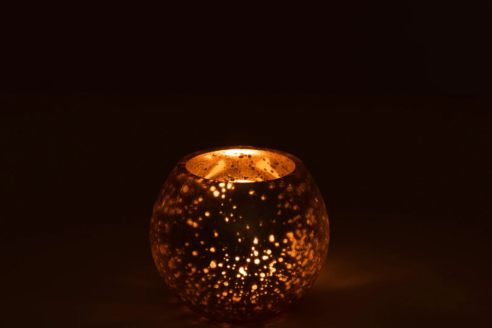 De Kaarsenhouder Orkaanbal Kraakglas Zilver Goud Klein gloeit warm in het donker, werpt een zacht gouden licht en sterachtige patronen op het gespikkelde, ronde oppervlak en de omliggende gebieden.
