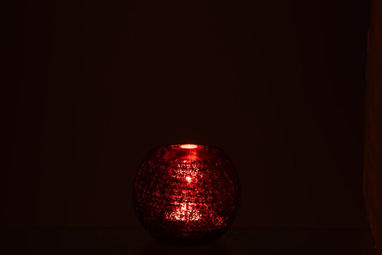 De Orkaanbal Knisperglas Glanzend Rood Klein, een kleine rood glazen kandelaar met textuur, gloeit zacht in het donker en werpt een warm rood licht. De zachte verlichting steekt af tegen de meestal zwarte achtergrond.