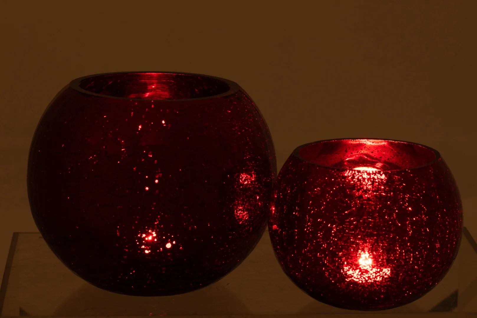 De Kaarsenhouder Orkaan Bal Kraakglas Rood Groot is een ronde, rode glazen kandelaar met een getextureerde afwerking. Hier afgebeeld naast een kleinere brandende houder, creëert hij een stijlvol display en werpt hij een warme gloed.