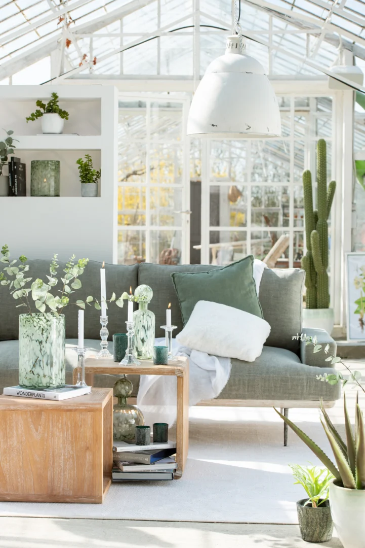 Een lichte leefruimte met een grijze bank, saliegroene kussens, houten tafels, groene planten en kaarsen is ingericht met de Kandelaar Klassiek Glas Transparant Groot als glasdecor in een zonovergoten serre met grote ramen en witte muren.
