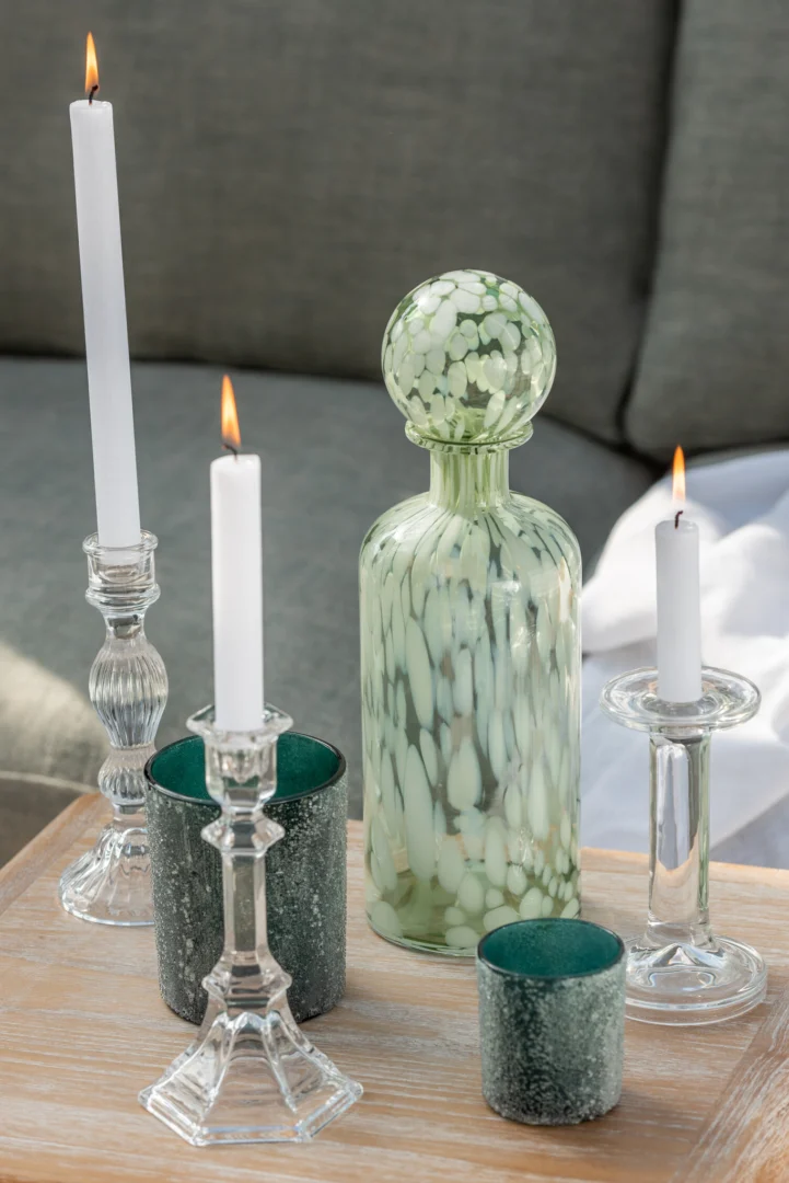 Op een houten dienblad staan drie brandende witte kaarsen in glazen houders, twee groene votiefhouders en de Kandelaar Klassiek Glas Transparant Groot decoratiefles met ronde stop, voor een grijze sofa.