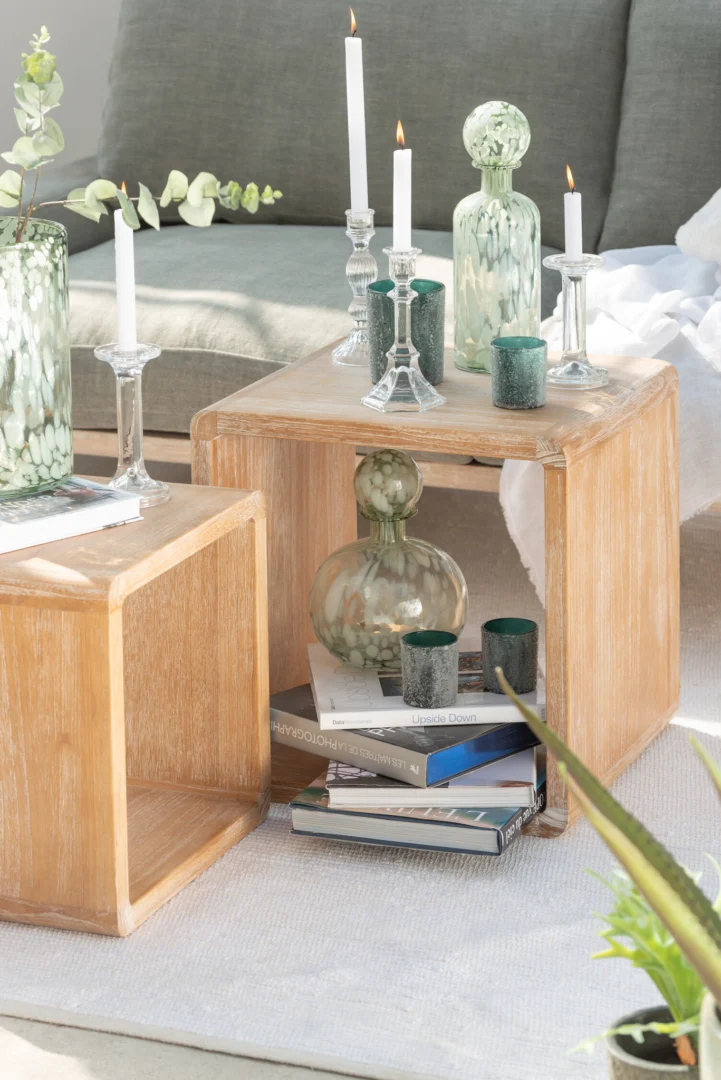 Op twee houten kubustafels staan groene glazen vazen, gestapelde boeken en witte kaarsen in de Kandelaar Klassiek Glas Transparant Groot houders. In één tafel staan een vaas en extra kaarsen. Op de achtergrond staan een grijze bank en een kleine plant.