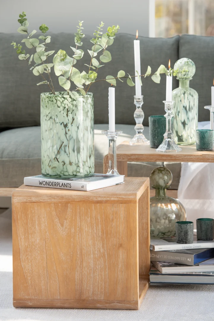 Op een houten kubustafel staan een groen gemarmerde vaas met eucalyptus en een boek met de titel WONDERPLANTEN. Op de achtergrond staan de Kandelaar Klassiek Glas Transparant Groot en decoratieve flessen op een andere houten tafel in een gezellige woonkamer.