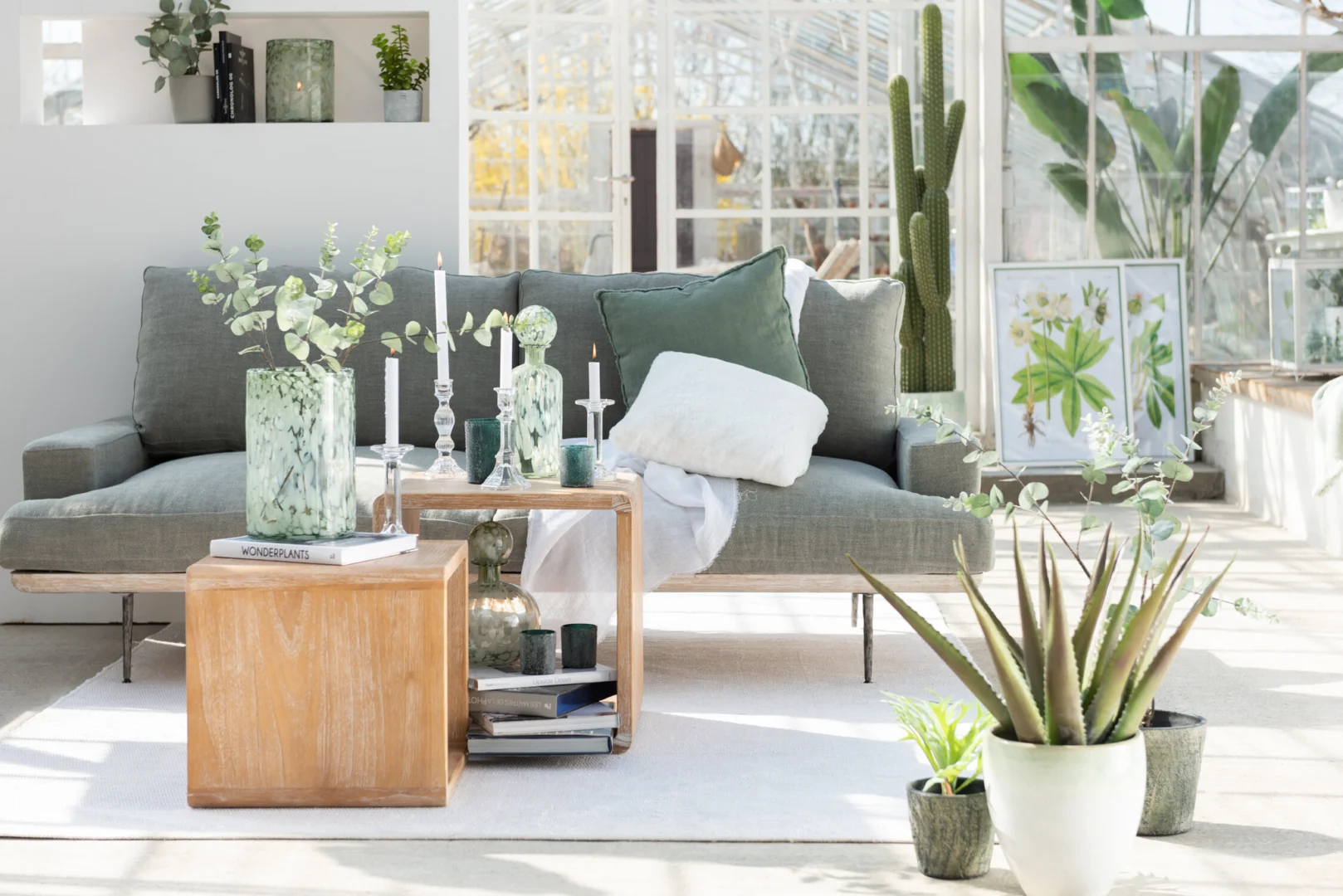 Een lichte, moderne woonkamer met een groene bank, een houten bijzettafel, potplanten, botanische prints en de Kandelaar Klassiek Glas Transparant Groot kandelaar in een zonovergoten ruimte met grote ramen.