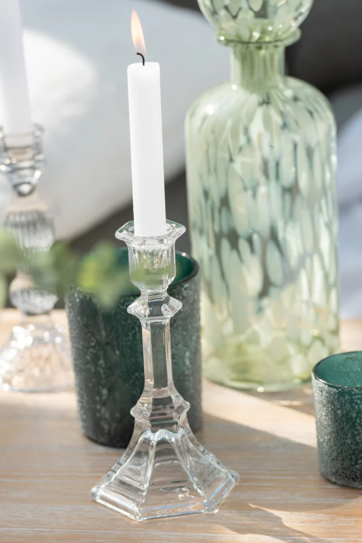 Een brandende witte kaars staat in de Kandelaar Klassiek Glas Transparant Groot op een houten tafel, omringd door groene glazen votiefhouders en een decoratieve groene glazen fles op de achtergrond.