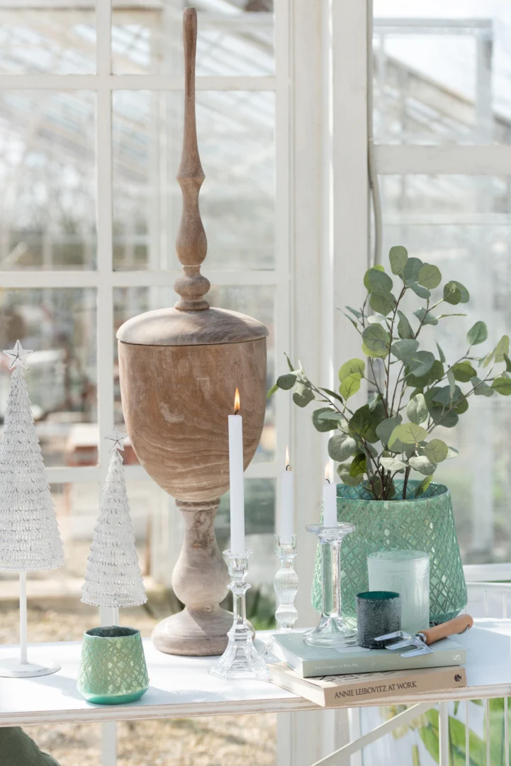 Een heldere binnenscène met een grote houten decoratieve urn, Kandelaar Klassiek Glas Transparant Groot die een witte kaars vasthoudt, miniatuur witte kerstboompjes, groene kaarsen, een bladplant in een groenblauwe mand en gestapelde boeken op een witte tafel.