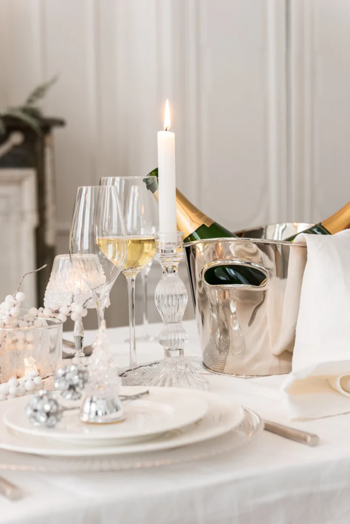 Elegante tafelschikking met witte borden, kristallen glazen, een Kandelaar Bal Glas Transparant Groot met een brandende kaars en een zilveren ijsemmer met champagne. Zilveren ornamenten en feestelijke decoraties dragen bij aan de verfijnde kerstsfeer.