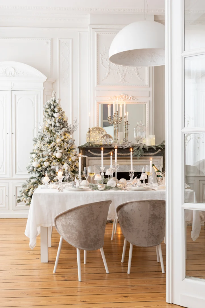 Een lichte, elegante eetkamer ingericht voor Kerstmis is voorzien van een wit tafelkleed, kaarsen, ornamenten en de Kandelaar Bal Glas Transparant Groot bij een berijpte boom bij de open haard, waardoor een gezellige kerstsfeer wordt gecreëerd.