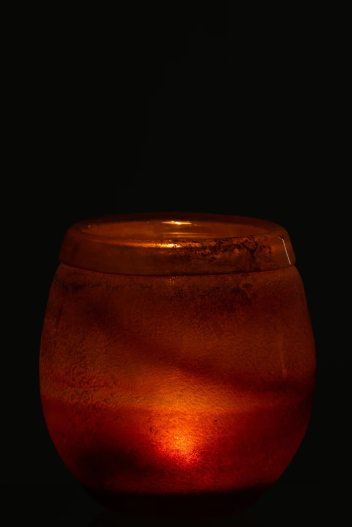 De Kaarsenhouder Orkaan Scavo Randglas Bourgondië Medium gloeit warm met een oranje kaars in de ronde matglazen houder, tegen een zwarte achtergrond.