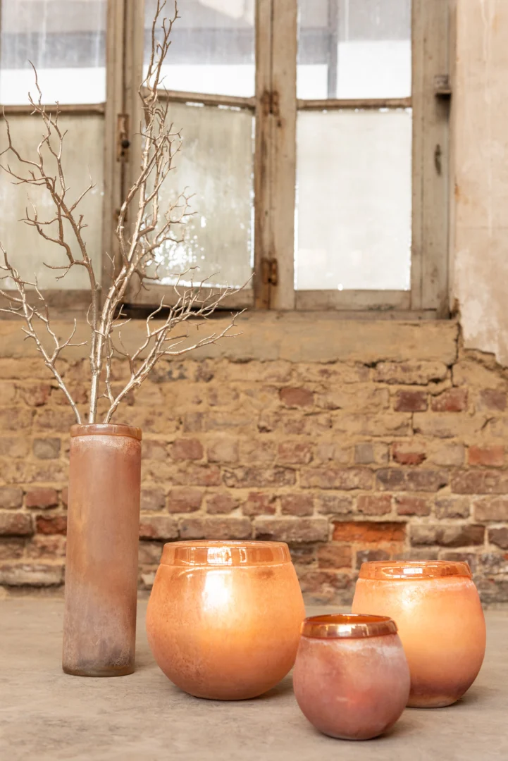 De Kaarsenhouder Orkaan Scavo Randglas Bourgondië Medium, een warmgetinte glazen kandelaar, staat op een betonnen vloer voor een verweerde bakstenen muur en oud houten raam, te glimmen tussen ronde glazen vazen.