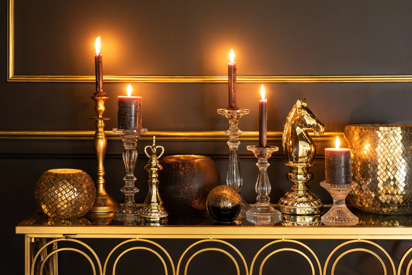 Een Kaarsenhouder Tibo Glas Transparant Groot met brandende kaarsen, gecombineerd met gouden accenten en een gouden paardenbeeldje, siert een gouden consoletafel tegen een donkere muur met gouden afwerking.
