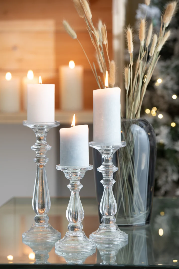 Drie witte pilaarkaarsen worden aangestoken en geplaatst op Kaarsenhouder Tibo Glas Transparant Groot kandelaars op een reflecterend glazen oppervlak, met decoratieve grassen in een heldere vaas en zacht gloeiende kaarsen op de achtergrond.