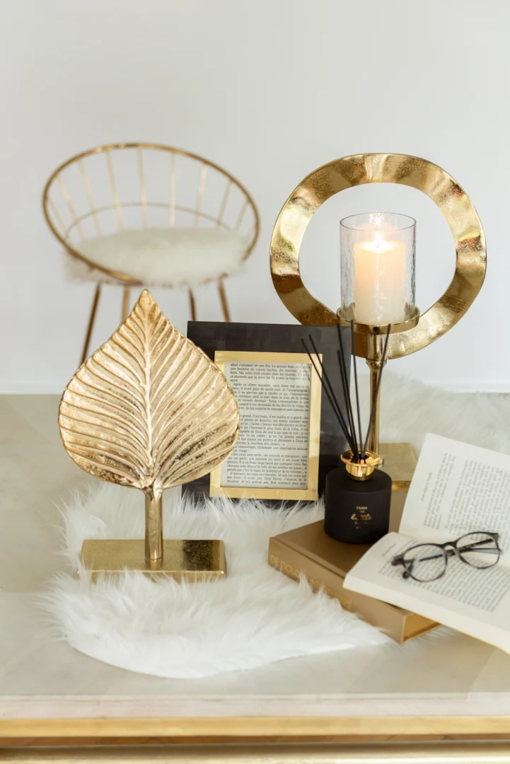 Een moderne decoropstelling met de Kaarsenhouder Orkaan Voet Aluminium Goud Klein kandelaar met een brandende kaars, een bladgouden ornament, een open boek met glazen, een rietverdamper en een wit bont vloerkleed; op de achtergrond verschijnt een gouden stoel.