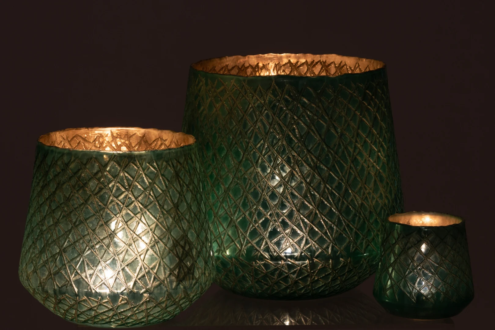 Drie Kaarsenhouder Orkaan Krassen Glas Mint Groen Groot kandelaars, elk met een brandende kaars erin, staan tegen een donkere achtergrond. Het mintgroene structuurglas creëert een warm, gloeiend effect.