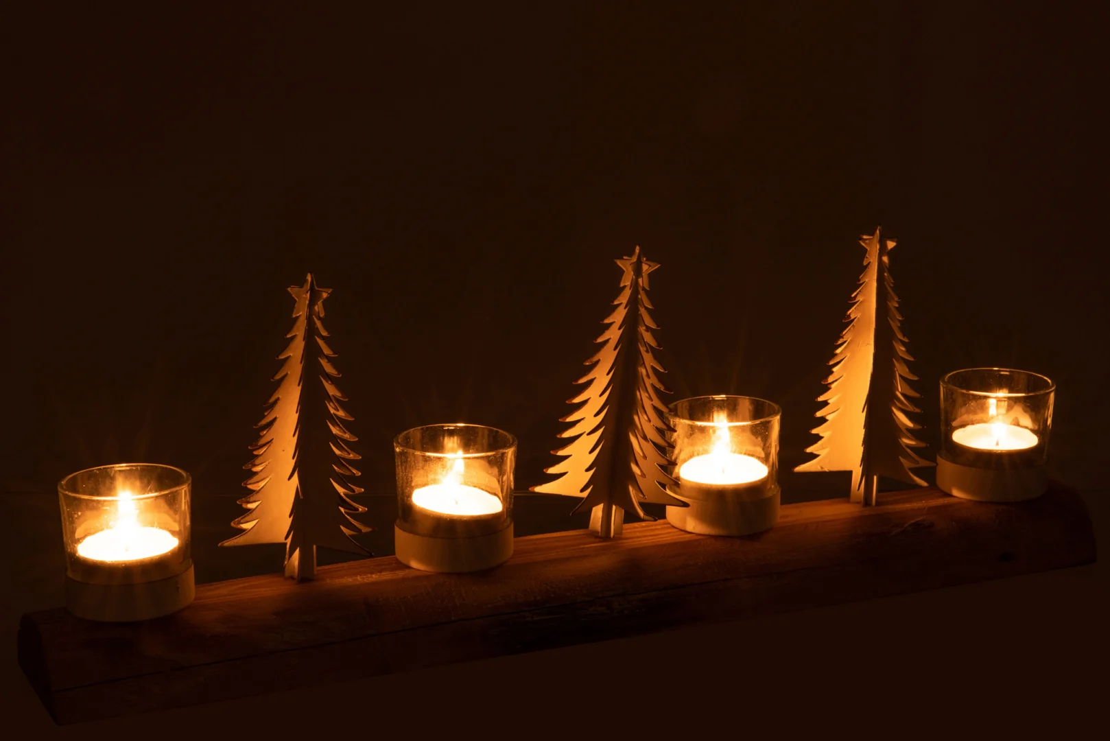De Theelichthouder Kerst Hout Metaal Wit XL heeft vier brandende theelichtjes in glazen houders op een houten voet, geaccentueerd door vier witte metalen kerstbomen, die een warme, feestelijke gloed werpen tegen een donkere achtergrond.