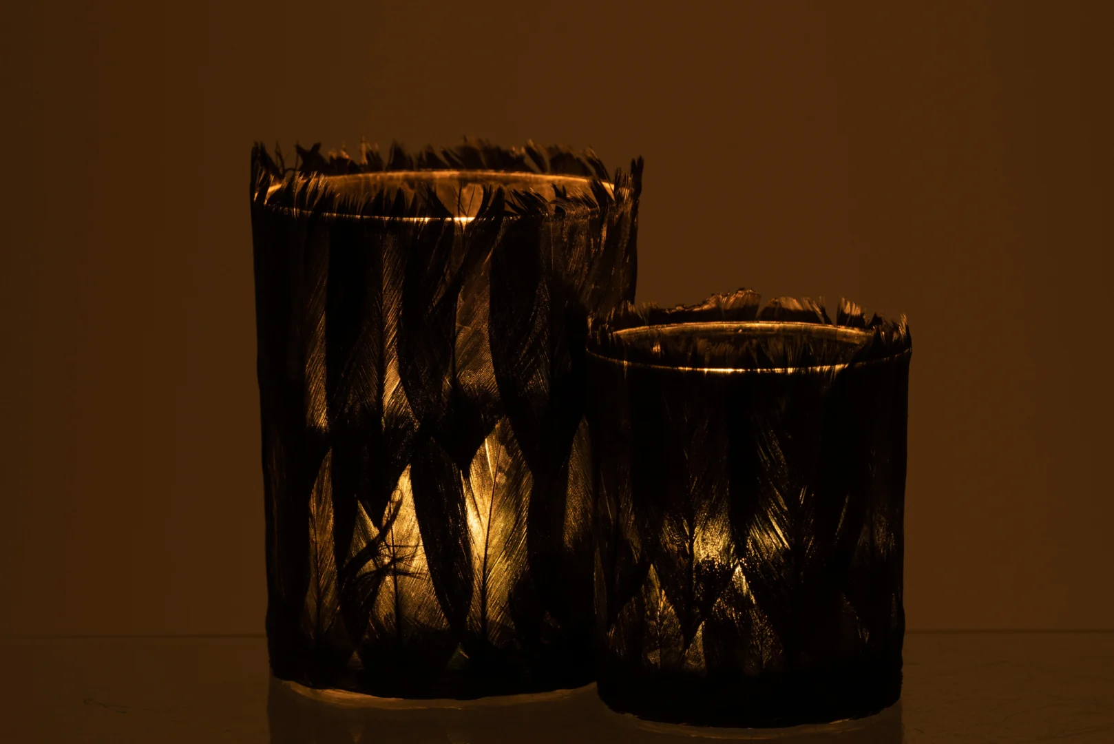 De Theelichthouder Veren Glas Zwart Groot is een hoge, decoratieve kandelaar met een donker, gestructureerd ontwerp, dat een warme gele gloed werpt in schemerig licht.