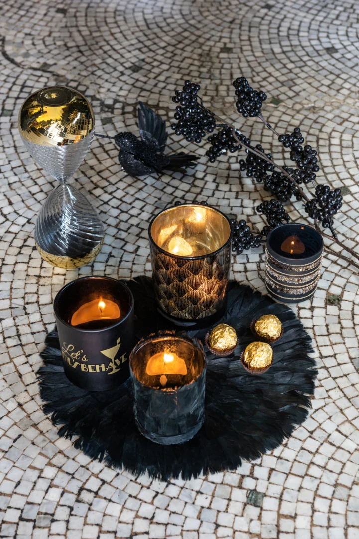 Een decoratieve display met drie verlichte Theelichthouder Veren Glas Zwart Groot kandelaars, een zandloper, zwarte kunstbessen, zwarte veren en in goud verpakte chocolaatjes op een oppervlak van mozaïektegels.