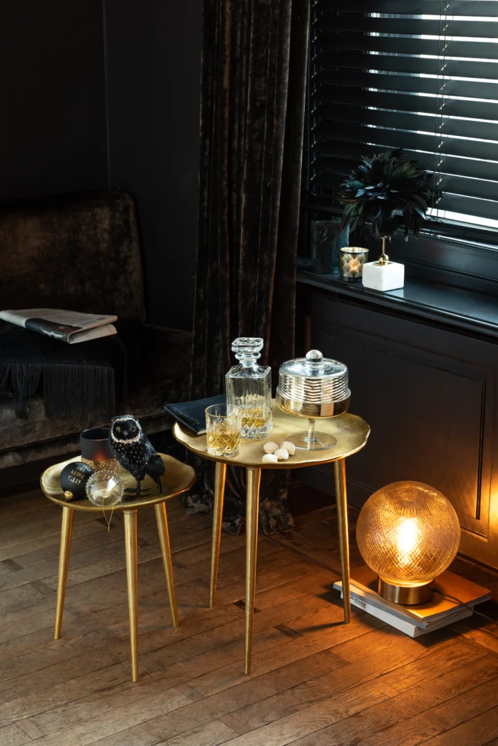 Een stijlvol interieur met twee ronde gouden tafels met glaswerk, een karaf, kaarsen, ornamenten en de Theelichthouder Veren Glas Zwart Groot. Een gloeiende lamp staat op de houten vloer met daarachter donkere gordijnen, zwarte jaloezieën en fluwelen stoelen.