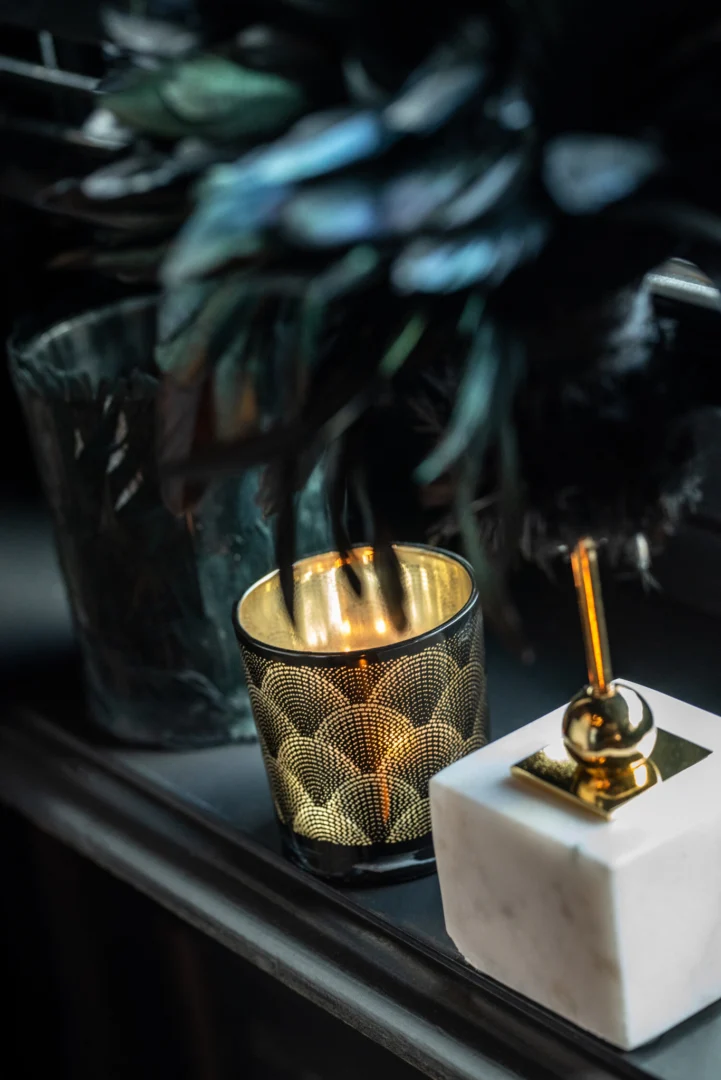De Theelichthouder Veren Glas Zwart Groot staat op een donker oppervlak naast een marmeren en gouden decorstuk, met vaag gebladerte op de achtergrond. Warm kaarslicht uit de houder creëert een gezellige sfeer.