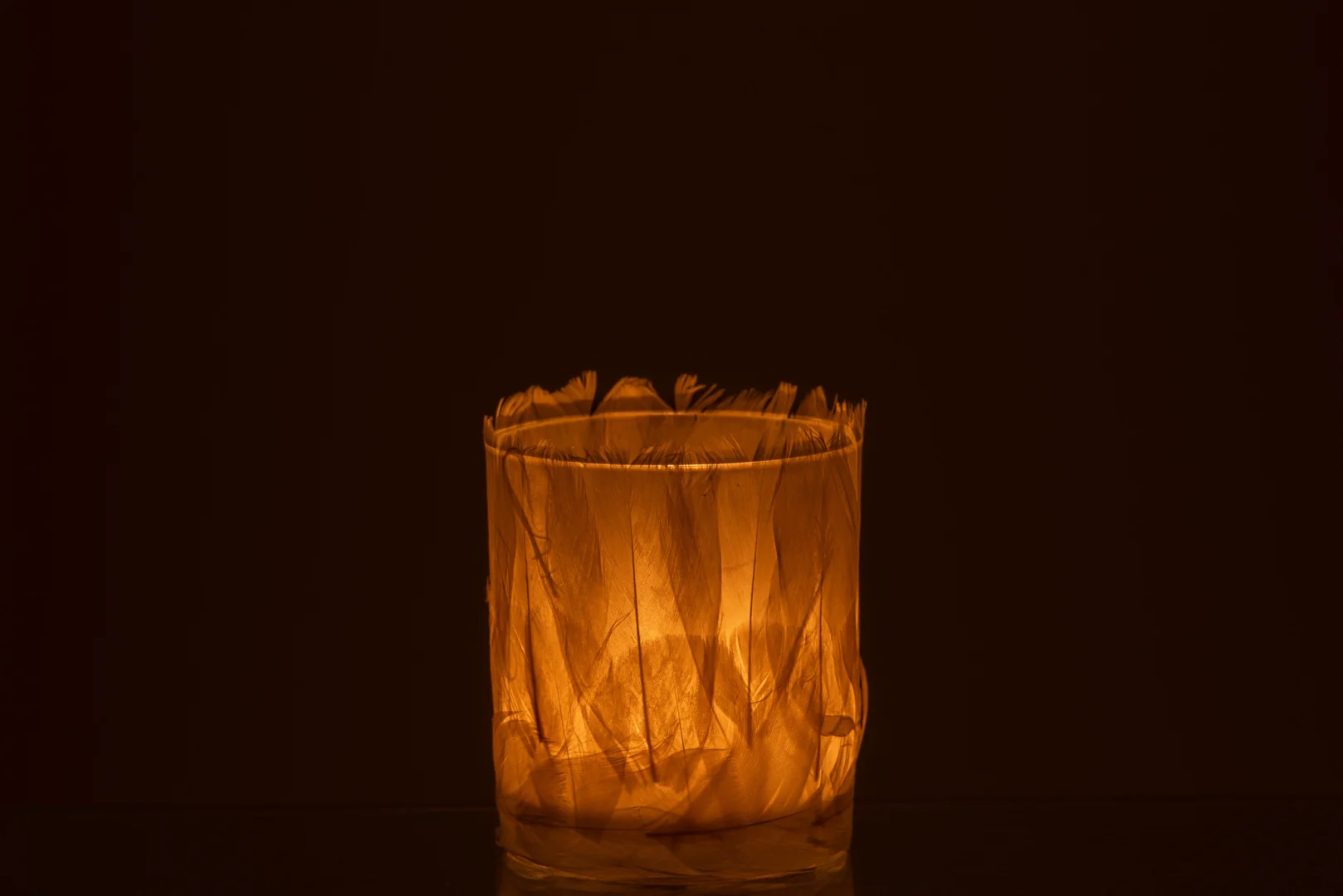 Een brandende kaars gloeit warm in de Theelichthouder Veren Glas Crème Klein, een kleine cilindervormige houder met delicate doorschijnende verenpatronen tegen een donkere achtergrond.