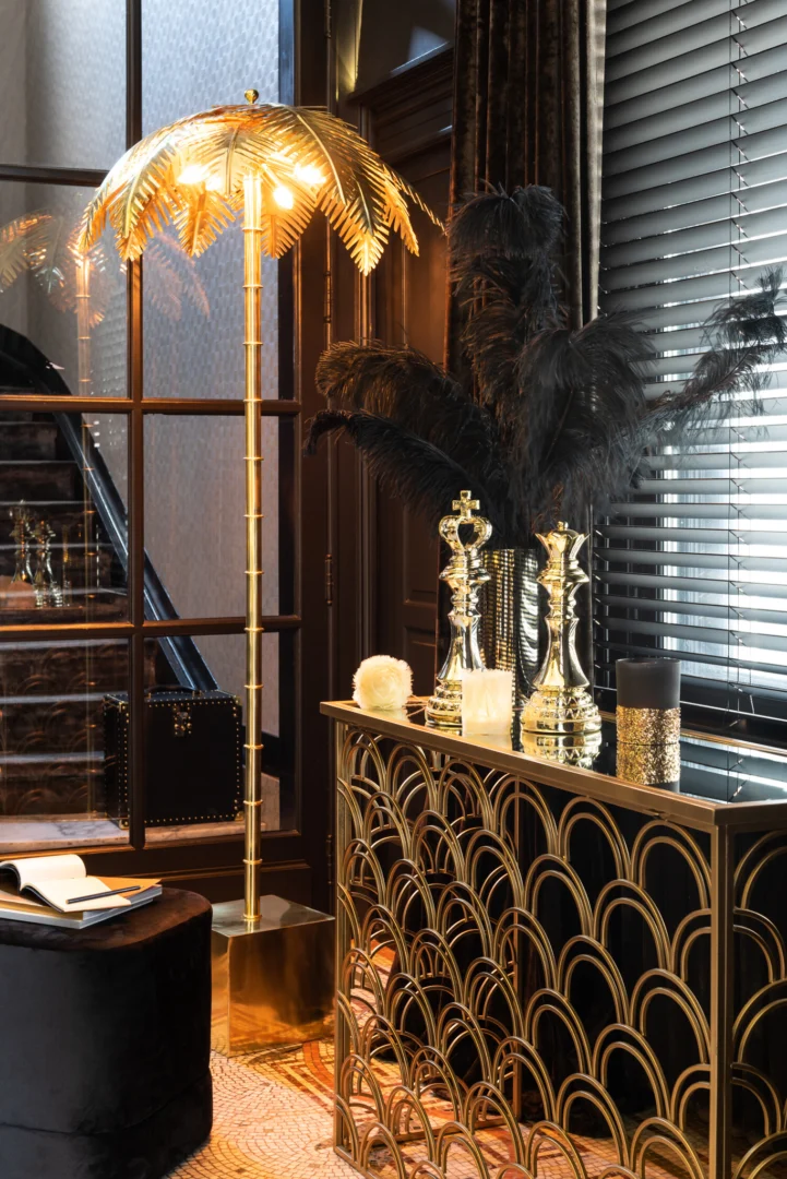Een stijlvol interieur met een gouden palmboom vloerlamp, art deco consoletafel, zwart verendecor, gouden schaakstukken, Theelichthouder Veren Glas Crème Klein en objecten van getextureerd glas bij een raam met jaloezieën en donkere gordijnen.