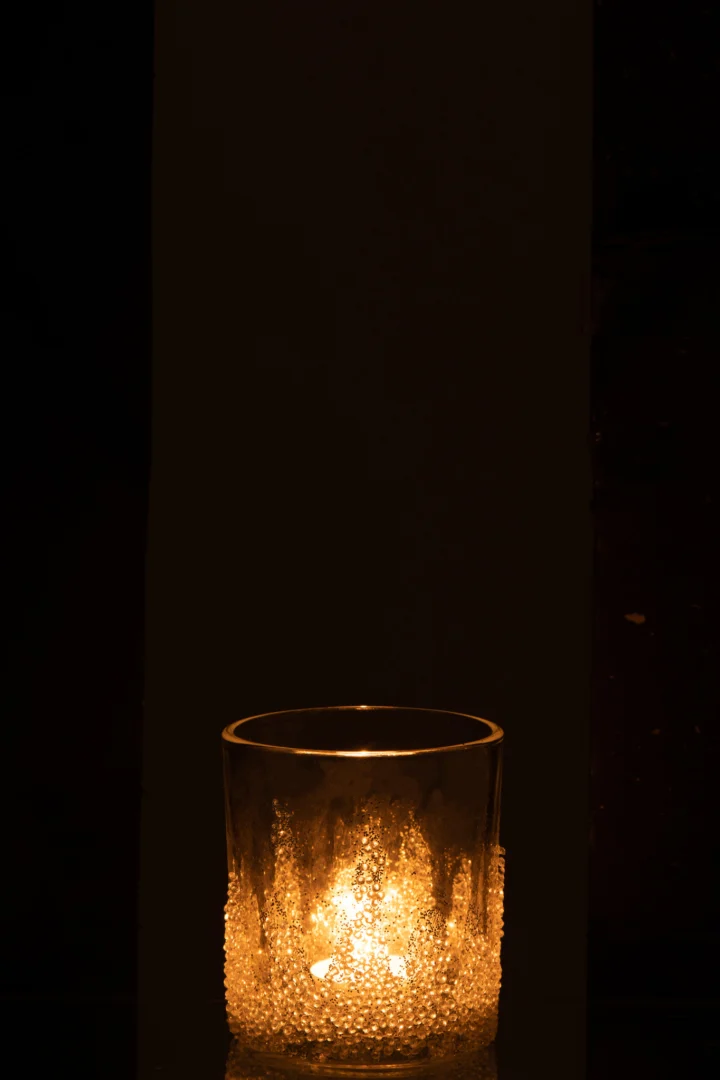 De Theelichthouder Suikerbodem Glas Helder Middel gloeit zacht in het donker, het getextureerde glas werpt een warm, gouden licht. Tegen een bijna zwarte achtergrond valt de zachte verlichting van de kaars prachtig op.