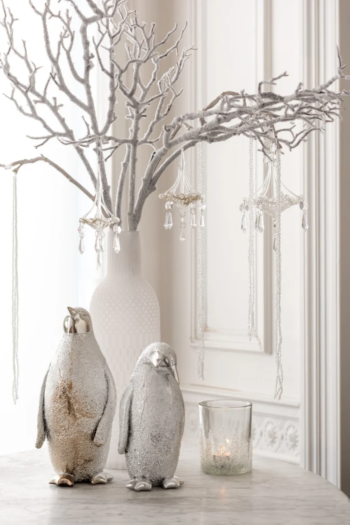 Een witte vaas met zilveren takken en hangende kristallen staat achter twee glinsterende pinguïnfiguurtjes, naast een kaarsje in de Theelichthouder Suikerbodem Glas Helder Middel op een marmeren oppervlak.