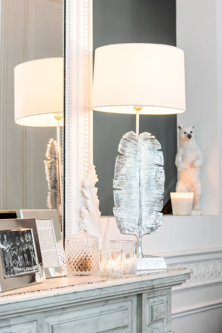 Een decoratieve schoorsteenmantel toont een zilveren verensculptuur, ingelijste foto's, een Theelichthouder Suikerbodem Glas Helder Middel kandelaar en een brandende lamp met een witte kap, met een wit berenbeeldje en een spiegel op de achtergrond.