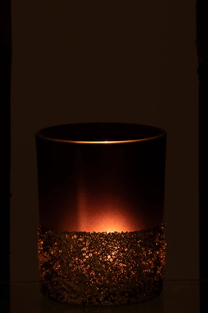 De Theelichthouder Pailletten Glas Blauw Goud Medium toont een brandende kaars in een donkere kamer, de metalen houder versierd met glitterende gouden lovertjes op de onderste helft, die een warme, zachte gloed verspreiden.