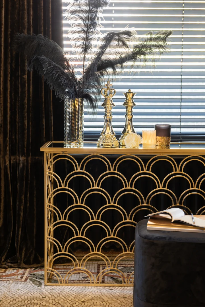 Een gouden consoletafel met een art-decopatroon toont hoge verenarrangementen in glazen vazen, sierlijke kandelaars en Theelichthouder Pailletten Glas Blauw Goud Medium. Jaloezieën bedekken het raam erachter en een boek ligt open op een nabijgelegen donker gestoffeerde kruk.