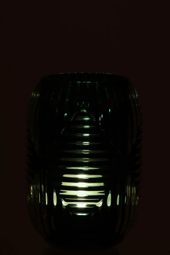 De Theelichthouder Hoge lijnen Glas Blauw heeft een gloeiende kaars in een geribbelde blauwe glazen houder, die zacht, gedessineerd licht werpt door de horizontale lijnen tegen een donkere achtergrond.