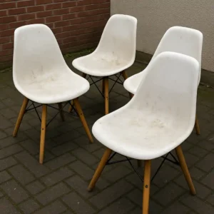Vier witte plastic stoelen met houten poten staan buiten op een bakstenen patio bij een bakstenen muur en een witte muur. De stoelen lijken wat vies-krijg je witte stoelen weer stralend schoon met een goede reiniging.