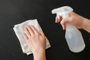 Iemand demonstreert hoe je matzwart hout schoonmaakt (hoe maak ik matzwart hout schoon) door een spuitfles in de ene hand te houden en het oppervlak af te vegen met een witte doek, zodat het vlek- en streeploos blijft.