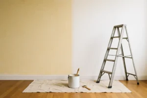 Een trapladder staat naast een muur die gedeeltelijk van beige naar wit is geschilderd om te bewijzen dat je vergeelde verf weer wit kunt krijgen. Een verfblik, kwast en hamer rusten op een droogdoek die de hardhouten vloer bedekt.