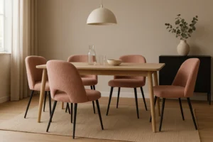 Een minimalistische eetkamer met een houten tafel, vijf oudroze eetkamerstoelen, een glazen karaf, twee glazen en een schaal op tafel. Erboven hangt een hanglamp en op de achtergrond staat een vaas met takken op een zwarte kast.