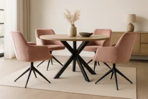 Een ronde houten eettafel met zwarte poten wordt omringd door vier roze eetkamerstoelen draaibaar op een licht vloerkleed. Een vaas met gedroogd pampagras en een schaal staan op tafel, terwijl een dressoir en een lamp op de achtergrond staan.