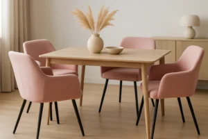Een minimale eethoek met een lichte houten tafel, vier roze eetkamerstoelen met armleuning, een vaas met gedroogd pampagras, een schaal en een lamp op een zijkast - alles in een zacht verlichte, neutraal getinte kamer.