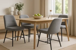 Een moderne eethoek met een lichte houten tafel, vier grijs gestoffeerde stoelen-waarom zijn grijze stoffen eetkamerstoelen zo geliefd?-een schaal met groene appels, een glazen karaf, twee glazen en beige gordijnen die natuurlijk licht binnenlaten.