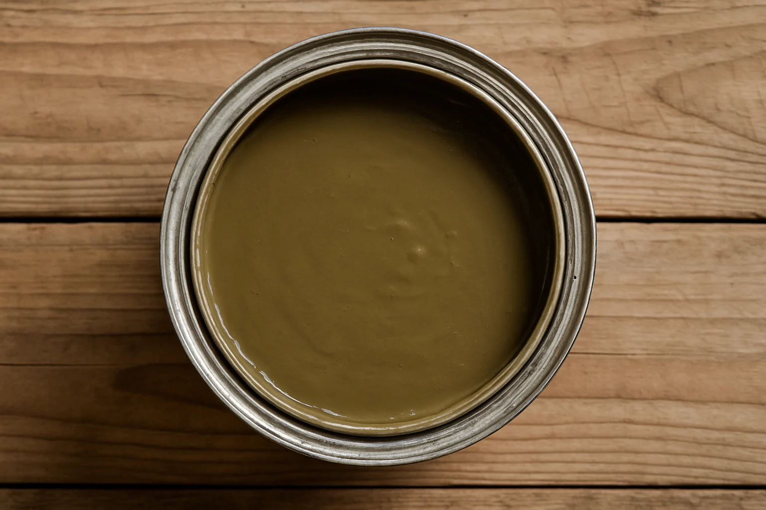 Een open blik gevuld met olijfgroene verf staat op een houten oppervlak, van bovenaf gezien. De verf lijkt glad en glanzend; sommigen zeggen dat het lijkt op "wat is de lelijkste kleur ter wereld," maar toch steekt het af tegen de natuurlijke houtnerfachtergrond.