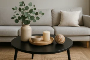 Een moderne woonkamer met een lichtgrijze bank en een ronde zwarte salontafel, perfect voor wie zich afvraagt "Wat zet ik op een zwarte salontafel?". De tafel is voorzien van een vaas met eucalyptus, kaarsenplateau, schaal en decoratieve bol.