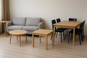 Een moderne woonkamer met een grijze bank, twee houten salontafels-maar welke tafel neemt de hoofdrol?-een kleine ronde bijzettafel en een houten eettafel met vier zwarte stoelen, allemaal op een lichthouten vloer tegen een effen beige muur.
