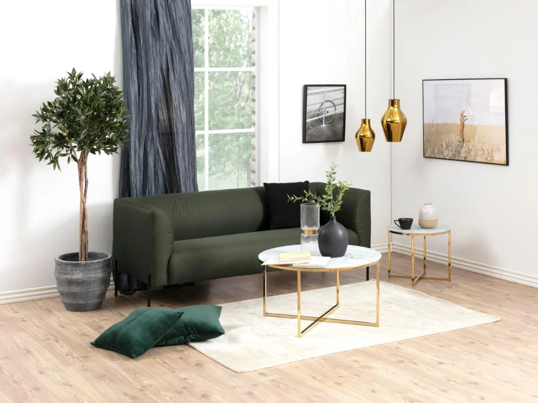 Een moderne woonkamer met een groene bank, boom in pot, wit vloerkleed, twee groene kussens, bijzettafeltjes met goudaccenten en een Salontafel 80 CM Rond Kunststof in Scandinavische stijl. Drie hanglampen en ingelijste foto's sieren de witte muren.